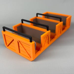 May include: An orange 3D-printed organiser with four compartments. Each compartment has a black handle and a brown interior. The sides of the organiser have a wooden crate design.