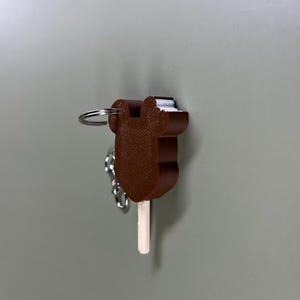 Pode incluir: Um chaveiro de sorvete de chocolate marrom com um palito branco e um anel de chave prateado.