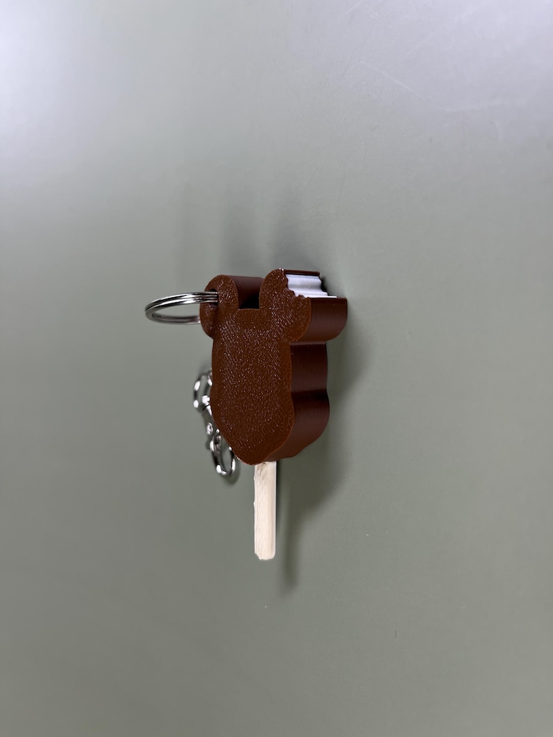 Pode incluir: Um chaveiro de sorvete de chocolate marrom com um palito branco e um anel de chave prateado.