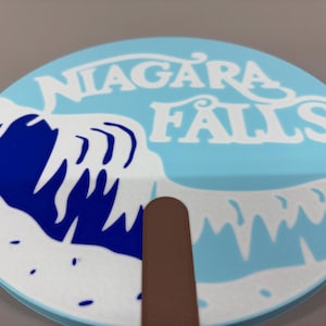 May include: A round hand fan featuring a light blue background with a white graphic of a crashing wave and the text NIAGARA FALLS in white lettering. The fan has a brown handle attached to the bottom center of the circular surface.