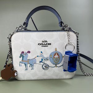 Pode incluir: Uma bolsa Coach branca com uma al&ccedil;a azul e uma al&ccedil;a de corrente prateada. A bolsa apresenta uma estampa de uma carruagem puxada por cavalos e um personagem de um filme da Disney. A bolsa tem um pequeno amuleto azul.