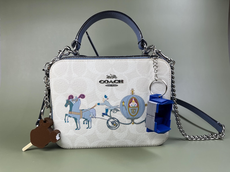 Pode incluir: Uma bolsa Coach branca com uma al&ccedil;a azul e uma al&ccedil;a de corrente prateada. A bolsa apresenta uma estampa de uma carruagem puxada por cavalos e um personagem de um filme da Disney. A bolsa tem um pequeno amuleto azul.