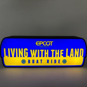 May include: A rectangular sign with a blue background and yellow text that reads "LIVING WITH THE LAND BOAT RIDE." The sign also includes the text "EPCOT PRESENTS" in yellow. The sign has a black border.