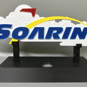 May include: A white, blue and yellow sign with the word "SOARIN'" on it. The sign is mounted on a black base and has a red aeroplane on top.