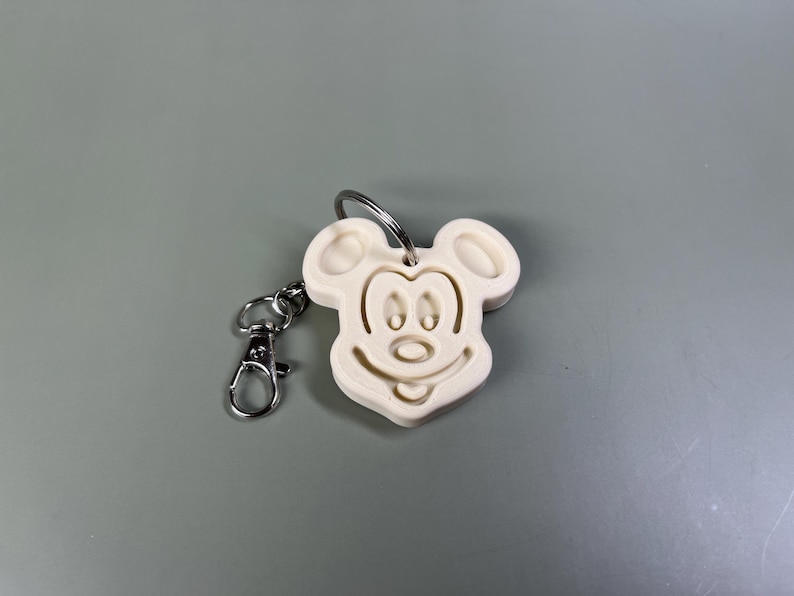 Pode incluir: Chaveiro de pl&aacute;stico branco com um design de cabe&ccedil;a do Mickey Mouse. O chaveiro tem um anel de metal prateado e um fecho de metal prateado.