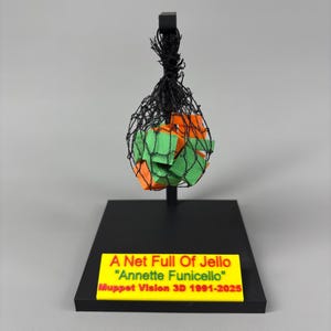 Puede incluir: Un soporte negro sostiene una red negra llena de formas geométricas naranjas y verdes. Un letrero amarillo en la base dice "A Net Full Of Jello" "Annette Funicello" Muppet Vision 3D 1991-2025.