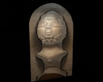 May include: A stone sculpture of a face with a crown, carved into a rectangular block. The sculpture is in a light brown color and has a textured surface.