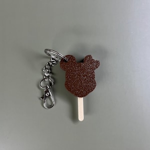 Pode incluir: Um chaveiro de chocolate marrom com a forma da cabe&ccedil;a do Mickey Mouse com um chaveiro prateado e um palito de madeira.