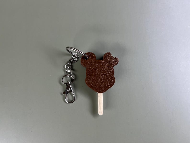 Pode incluir: Um chaveiro de chocolate marrom com a forma da cabe&ccedil;a do Mickey Mouse com um chaveiro prateado e um palito de madeira.