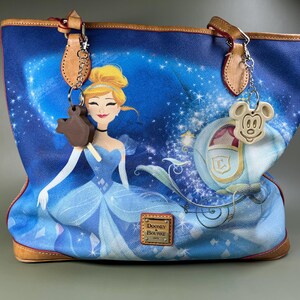 Pode incluir: Uma bolsa tote azul e marrom com uma estampa de Cinderela e sua carruagem. A bolsa tem o logotipo Dooney & Bourke e dois pingentes, um do Mickey Mouse e outro de uma barra de chocolate.