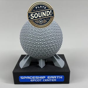 May include: A gray geodesic sphere model on a black rectangular base with a blue plaque reading SPACESHIP EARTH EPCOT CENTER. A circular gold and black emblem at the top features the text PLAYS SOUND! with musical notes and a speaker icon.