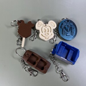 Pode incluir: Cinco chaveiros com designs diferentes. Um &eacute; uma barra de chocolate marrom, um &eacute; uma cabe&ccedil;a branca do Mickey Mouse, um &eacute; um carro azul, um &eacute; uma barra de chocolate marrom com uma fechadura e um &eacute; um chaveiro redondo azul com um design.