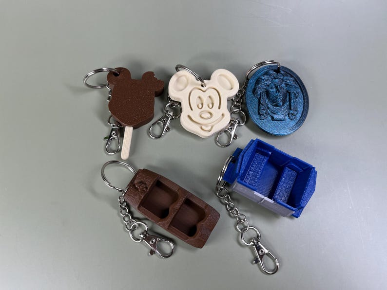 Pode incluir: Cinco chaveiros com designs diferentes. Um &eacute; uma barra de chocolate marrom, um &eacute; uma cabe&ccedil;a branca do Mickey Mouse, um &eacute; um carro azul, um &eacute; uma barra de chocolate marrom com uma fechadura e um &eacute; um chaveiro redondo azul com um design.