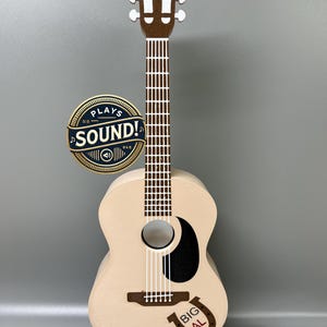 Könnte beinhalten: Eine hölzerne Akustikgitarre mit braunem Hals und weißen Stimmwirbeln. Der Korpus der Gitarre ist cremefarben mit einem schwarzen Schlagbrett und einem Hufeisen mit der Aufschrift "BIG AL". Die Gitarre steht auf einem schwarzen Ständer. Ein gold-schwarzer runder Aufkleber mit der Aufschrift "PLAYS THE SOUND!" befindet sich auf der linken Seite der Gitarre.