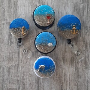 May include: Six different blue and white resin badge reels with various charms. The charms include a wine glass, an anchor, a fish, and a dolphin. The reels are attached to a clear plastic retractable cord.
