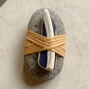 Op de afbeelding: Een grijze steen met een blauw-wit gestreept accent, omwikkeld met geweven bruin materiaal. De steen heeft een gladde, afgeronde vorm en staat tegen een neutrale achtergrond.