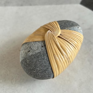 May include: A smooth, gray stone wrapped with woven, light brown bamboo. The bamboo is intricately woven around the stone, creating a decorative and natural aesthetic. The stone has a rounded, oval shape.