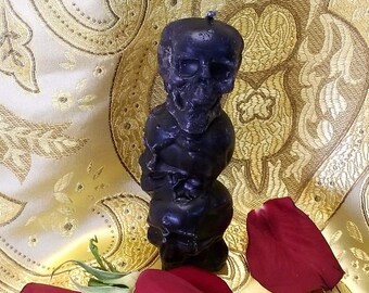 May include: A black candle shaped like three stacked skulls, sitting on a gold fabric background with red rose petals.