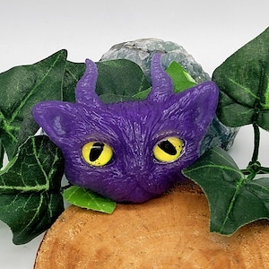 May include: Purple cat figurine with yellow eyes and horns. The cat is sitting on a wooden surface with green leaves.