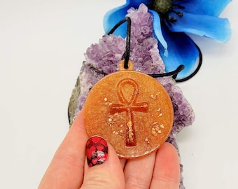 May include: A gold-toned pendant with an Ankh symbol, set in a round resin base with gold flecks. The pendant is on a black cord and is resting on a purple amethyst crystal.