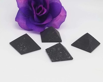 May include: Four black obsidian pyramids, each with a slightly different shape and size, are arranged on a white surface. A purple flower is out of focus in the background.
