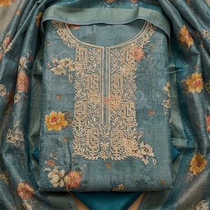 May include: A blue-grey fabric garment featuring a floral print with yellow and pink flowers. The neckline and front panel are decorated with intricate gold-toned embroidery and beadwork. A matching floral-patterned shawl drapes around the piece.