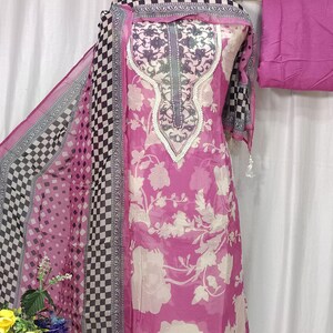 May include: A pink and white floral print salwar kameez set featuring a long tunic with a decorative neckline, a matching dupatta with a black and white checkered border, and a solid pink fabric piece, all displayed on a mannequin form.
