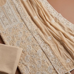 Op de afbeelding: Een beige driedelige kledingset bestaande uit een tuniek met witte bloemen- en paisley-borduursels, een bijpassend gevouwen beige stofstuk en een doorschijnende beige dupatta met wit geborduurde randen en kleine bloemmotieven.