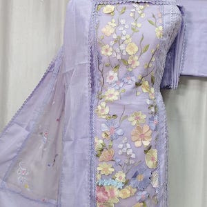 May include: A light purple embroidered salwar kameez set. The ensemble features a tunic, pants, and a scarf. The tunic is adorned with colorful floral embroidery and lace trim. The outfit is displayed on a mannequin.