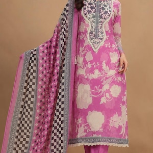 May include: A bright pink long-sleeved tunic featuring a large cream floral print, a dark grey patterned neckline, and a matching pink straight-leg pant. A long, sheer scarf with a black, white, and pink checkered border drapes over one shoulder.
