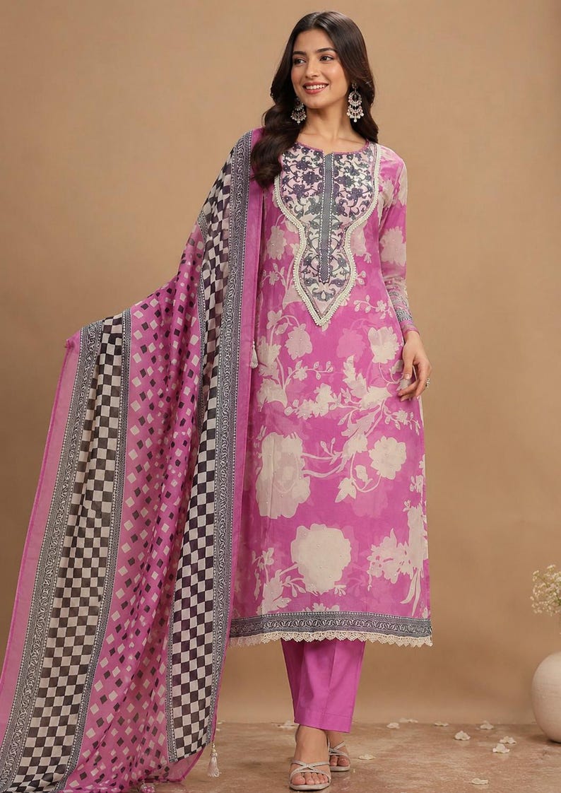 May include: A bright pink long-sleeved tunic featuring a large cream floral print, a dark grey patterned neckline, and a matching pink straight-leg pant. A long, sheer scarf with a black, white, and pink checkered border drapes over one shoulder.