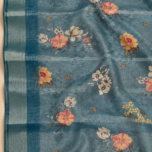 May include: A teal blue fabric featuring a floral pattern with pink, yellow, and white flowers. The textile has a subtle mottled texture and a wide vertical border on the left side with a metallic silver stripe running through the weave.