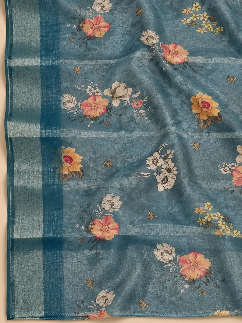 May include: A teal blue fabric featuring a floral pattern with pink, yellow, and white flowers. The textile has a subtle mottled texture and a wide vertical border on the left side with a metallic silver stripe running through the weave.