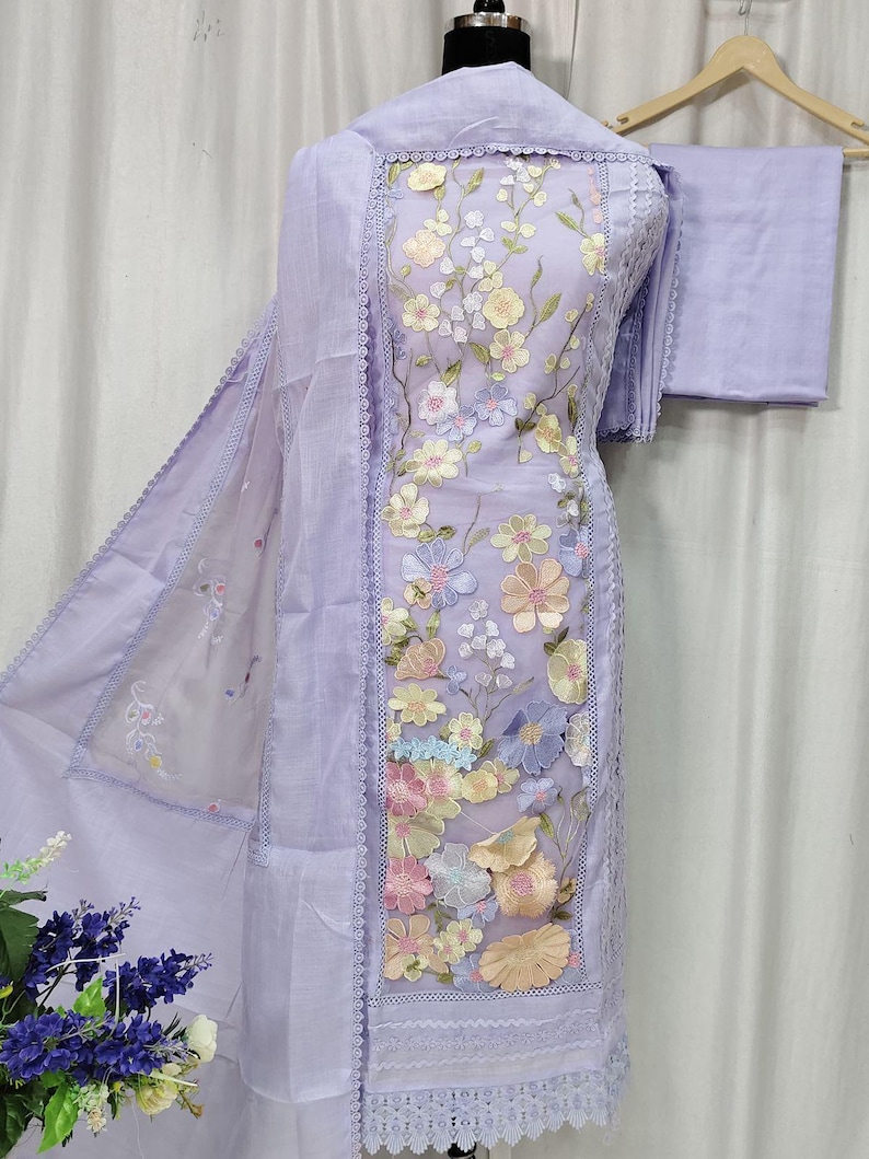 May include: A light purple embroidered salwar kameez set. The ensemble features a tunic, pants, and a scarf. The tunic is adorned with colorful floral embroidery and lace trim. The outfit is displayed on a mannequin.