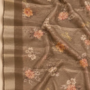 May include: A taupe-colored fabric featuring a repeating floral pattern with peach, orange, and white blossoms. The textile has a visible woven texture and a wide, metallic gold border along one edge, characterized by horizontal stripes.
