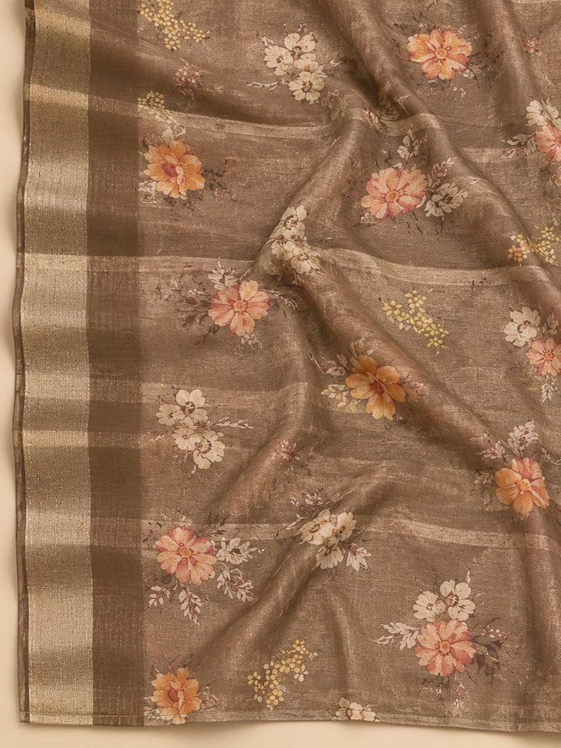 May include: A taupe-colored fabric featuring a repeating floral pattern with peach, orange, and white blossoms. The textile has a visible woven texture and a wide, metallic gold border along one edge, characterized by horizontal stripes.