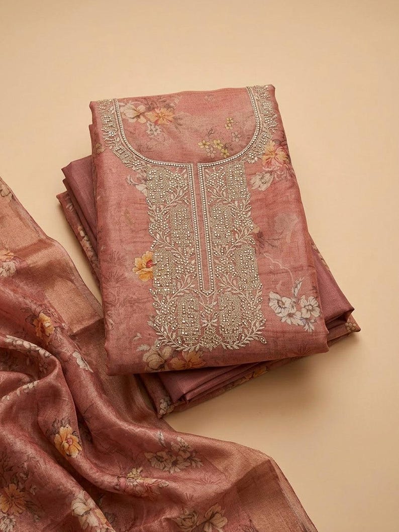 May include: A folded dusty rose fabric garment featuring a floral print in shades of yellow and white. The neckline and front panel are decorated with intricate silver embroidery and small metallic beads, paired with a matching floral fabric piece.