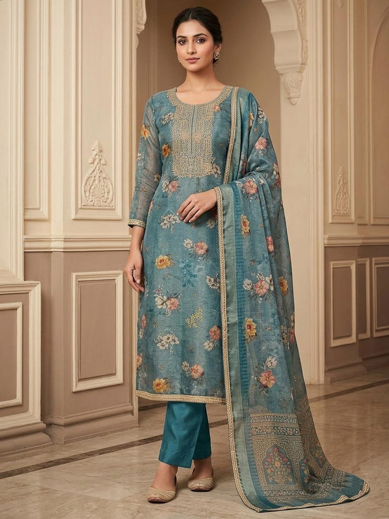 May include: A blue floral print long-sleeved tunic with a round neckline featuring gold embroidery, paired with solid teal trousers and a matching blue floral print dupatta with a gold border. The ensemble is crafted from a sheer, textured fabric.