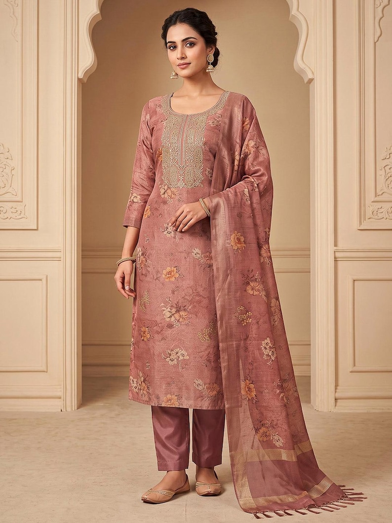 May include: A dusty rose three-piece ensemble featuring a knee-length tunic with three-quarter sleeves, floral patterns, and intricate gold embroidery around the neckline, paired with matching solid trousers and a coordinating floral print dupatta.