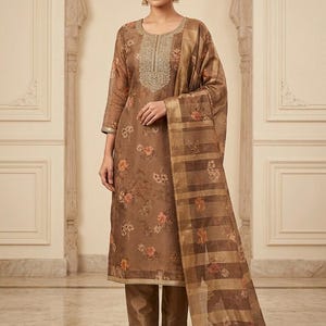 May include: A brown three-piece ensemble consisting of a long-sleeved straight-cut tunic with a floral print and gold embroidery around the neckline, matching solid brown trousers, and a sheer brown dupatta featuring a plaid pattern and floral motifs.