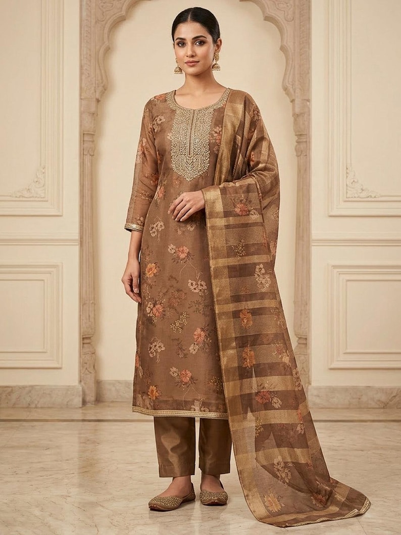May include: A brown three-piece ensemble consisting of a long-sleeved straight-cut tunic with a floral print and gold embroidery around the neckline, matching solid brown trousers, and a sheer brown dupatta featuring a plaid pattern and floral motifs.