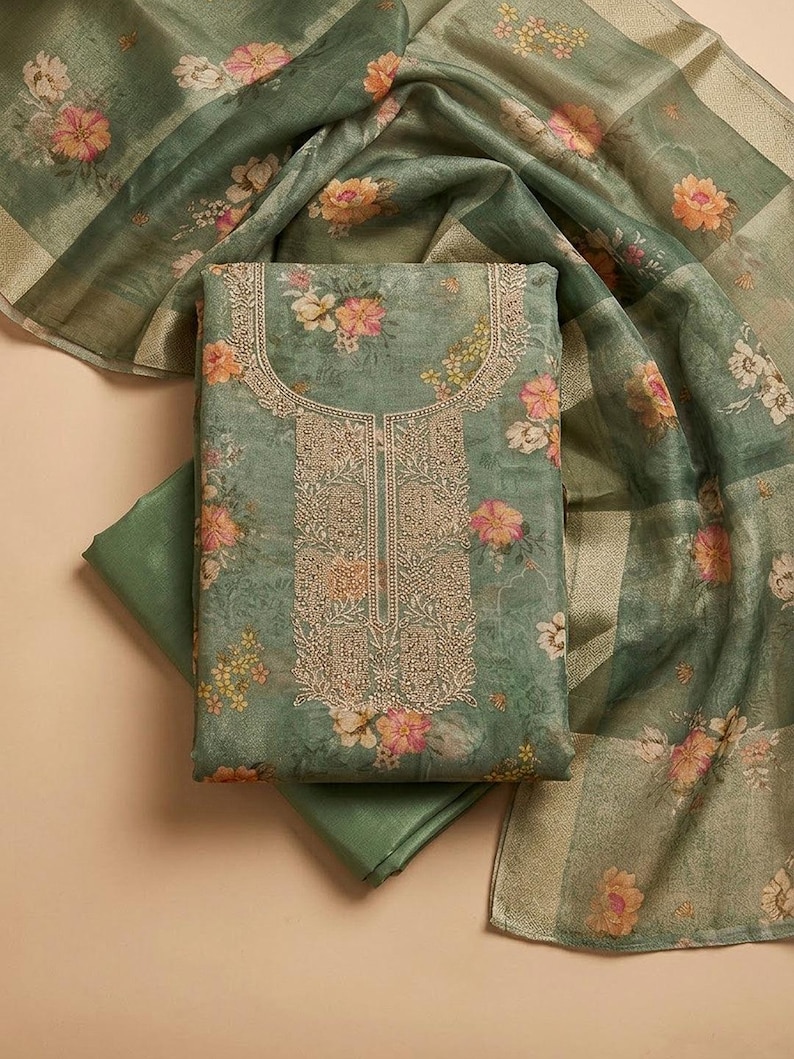 May include: A sage green fabric set featuring a floral print with pink and yellow blossoms. The central piece includes an intricate gold-toned embroidered neckline and front panel. A matching sheer, lightweight scarf with a floral pattern is included.