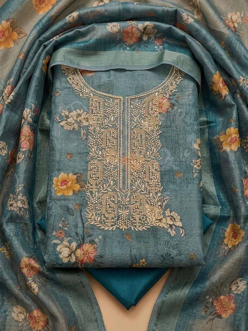 May include: A blue-grey fabric garment featuring a floral print with yellow and pink flowers. The neckline and front panel are decorated with intricate gold-toned embroidery and beadwork. A matching floral-patterned shawl drapes around the piece.