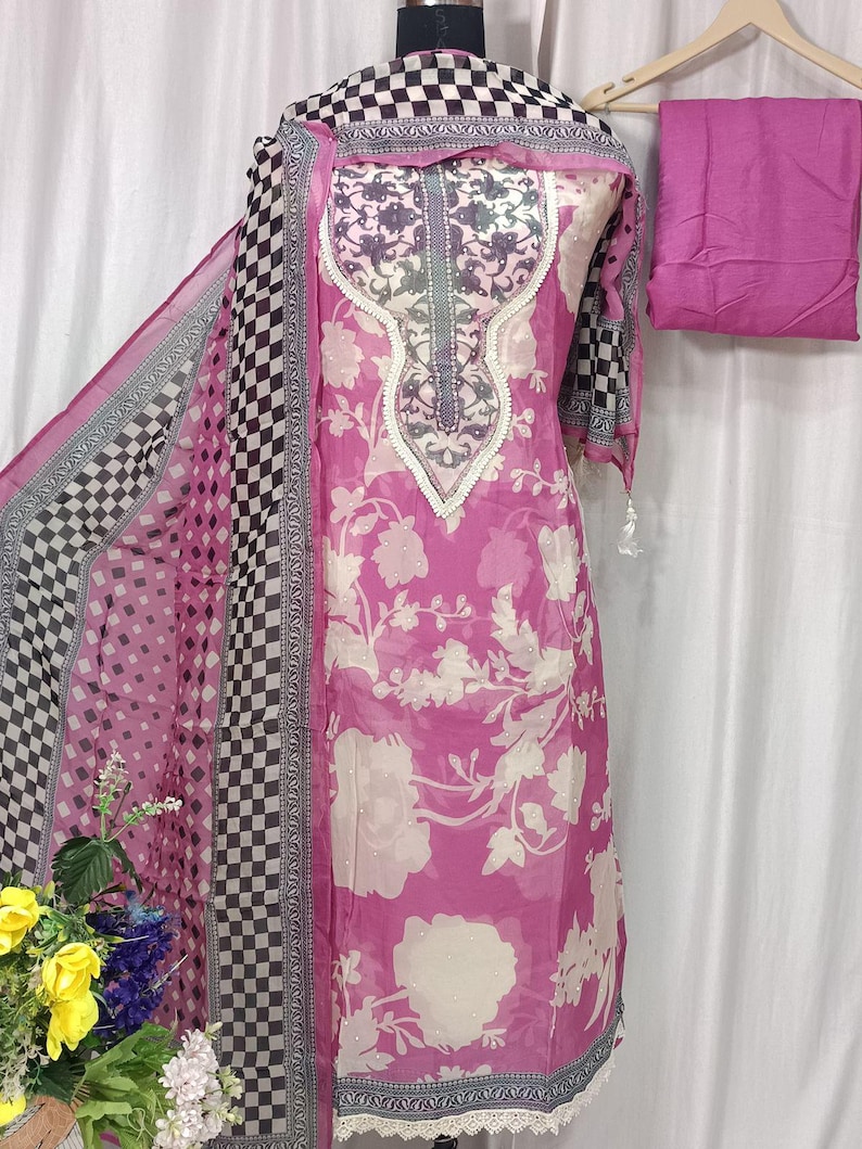 May include: A pink and white floral print salwar kameez set featuring a long tunic with a decorative neckline, a matching dupatta with a black and white checkered border, and a solid pink fabric piece, all displayed on a mannequin form.