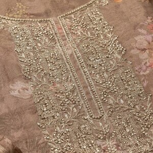 May include: A taupe-colored fabric garment featuring a floral print in muted tones of pink and orange. The neckline and front placket are heavily embellished with intricate silver-toned metallic embroidery, beadwork, and sequins in a floral pattern.