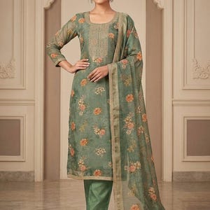 May include: A sage green salwar kameez set featuring a knee-length tunic with a scoop neckline, intricate gold embroidery, and a floral print. The outfit includes matching solid green trousers and a sheer, floral-patterned dupatta with gold trim.