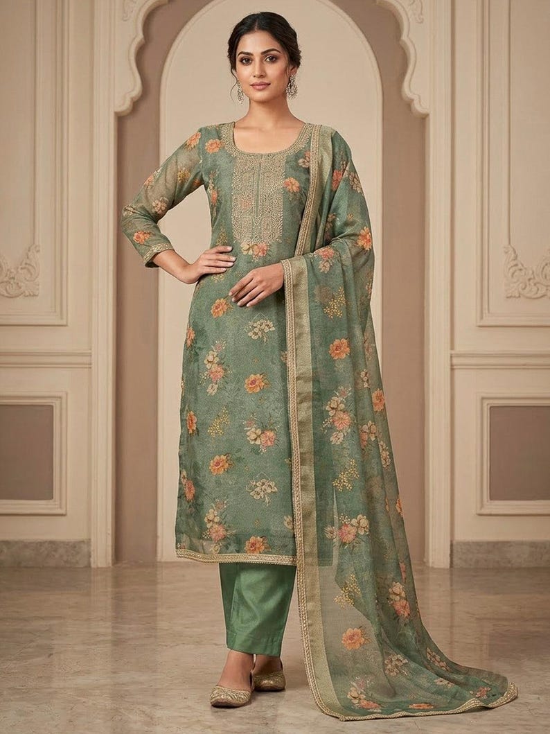 May include: A sage green salwar kameez set featuring a knee-length tunic with a scoop neckline, intricate gold embroidery, and a floral print. The outfit includes matching solid green trousers and a sheer, floral-patterned dupatta with gold trim.