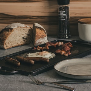 May include: A black cast iron griddle with a fried egg, bacon, and sausages. There is also a plate and a fork on the table.
