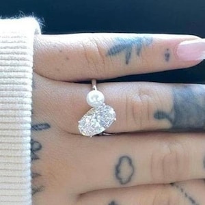 May include: A silver ring with a large oval-shaped clear stone and a smaller pearl on the side. The ring is on a person's finger with several tattoos.
