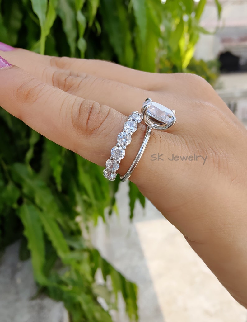 May include: A silver ring set with a large oval-shaped clear stone and a smaller band of clear stones. The ring is on a finger.